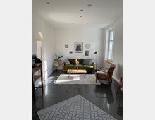 Helles Wohnzimmer mit weiß gestrichenen Wänden, Fliesenboden und einem großen Fenster rechts. Im Raum stehen ein grünes Sofa, ein Beistelltisch mit einer Vase, ein gemusterter Sessel und eine Stehlampe; an der Wand hängen Bilder und eine Uhr.