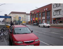 Straßenszene an der Hermannstraße in Berlin mit mehrstöckigen Wohngebäuden im Hintergrund, einem Einkaufszentrum rechts mit Geschäften wie REWE und Netto, sowie geparkten Autos, Fahrrädern und einer U-Bahn-Station (U8). Wetter ist bewölkt und nass, und Fahrzeuge fahren auf der Straße.