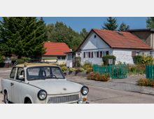 Ein weißes Retro-Auto steht in einer ruhigen Wohnstraße vor einem kleinen, weiß gestrichenen Haus mit roten Dachziegeln. Im Vorgarten des Hauses befinden sich Blumenbeete, ein grüner Zaun und Sträucher, umgeben von weiteren Häusern und Bäumen im Hintergrund.