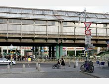Straßenszene unter einer erhöhten U-Bahn-Brücke aus Metall mit Glasfenstern. Im Vordergrund sind Fußgänger, Fahrräder und Straßenschilder, im Hintergrund Geschäfte, ein Fitnessstudio und geparkte Fahrräder sichtbar.