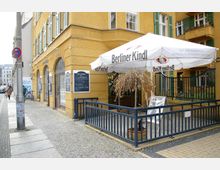Straßenszene mit einem gelben Gebäude im Eckstil, das ein Bistro beherbergt. Vor dem Eingang steht ein großer Sonnenschirm mit der Aufschrift „Berliner Kindl“ und eine kleine Terrasse mit Metallgeländer sowie einer Tafel mit dem Tagesangebot.