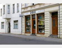 Straßenansicht einer Bäckerei mit der Aufschrift „KOLBO“ und „Barches Bakery“ auf dem Schaufenster. Das Gebäude hat eine helle Fassade mit dekorativen Verzierungen und Holzelementen an den Türen sowie große Glasfenster mit ausgestellten Produkten und blauen Davidsternen.