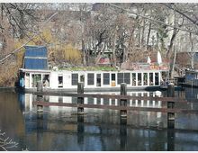 Hausboot auf einem ruhigen Fluss, umgeben von kahlen Bäumen im Winter. Das Boot hat ein bepflanztes Dach, große Fenster und eine Terrasse mit Sitzgelegenheiten; im Vordergrund sind Holzgeländer über dem Wasser zu sehen.