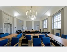 Ein heller Konferenzraum mit großen Fenstern, hohen Decken und einem modernen Kronleuchter. Der Raum ist mit Tischen und Stühlen in Blau und Schwarz ausgestattet, und im Hintergrund stehen Flipcharts und eine weiße Tafel.