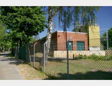 Eingezäuntes Gebäude mit roter Klinkerfassade und gelbem Anbau, umgeben von Bäumen und gelegen an einem Gehweg. Im Vordergrund ein Metallgitterzaun mit Stacheldraht, im Hintergrund technische Anlagen auf dem Dach.