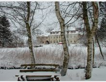 Schneebedeckte Parklandschaft mit einer Holzbank im Vordergrund, umgeben von schneebedeckten Birken. Im Hintergrund ein helles, historisches Gebäude mit mehreren Fenstern und einem Eingangstor, eingebettet in eine winterliche Szene.