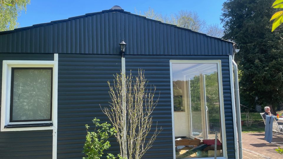 Dunkelblaues, eingeschossiges Holzhaus mit Satteldach, einem kleinen Fenster links und einem großen Fenster rechts, das als offene Tür wirkt. Davor stehen Sträucher, rechts schließt eine gepflasterte Terrasse an; im Hintergrund sind Bäume und ein klarer Himmel zu sehen.