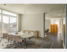 Heller Besprechungsraum mit einem ovalen Tisch, umgeben von modernen Bürostühlen auf einem Teppich. Große Fenster mit Jalousien lassen Tageslicht herein, und im Hintergrund ist ein Bereich mit Sesseln und einem gelben Raumteiler sichtbar.