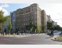 Straßenszene in einer Stadt mit einem mehrstöckigen Wohn- und Geschäftshaus aus beigem Putz im Vordergrund, umgeben von grüner Kletterpflanzenfassade. Im unteren Bereich des Gebäudes befinden sich ein Café mit Außensitzplätzen und Geschäfte, während auf der gepflasterten Straße Fahrradfahrer und parkende Autos zu sehen sind.