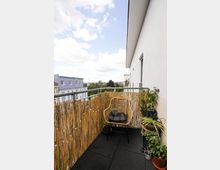 Ein kleiner Balkon einer Wohnung mit Bambus-Sichtschutz, einer Lichterkette, einem Korbstuhl mit Sitzkissen und mehreren Pflanzen in Töpfen. Im Hintergrund sind Wohngebäude und ein bewölkter Himmel zu sehen.