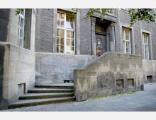 Historische Steinfassade eines Gebäudes mit hohen Fenstern und einer breiten Treppe, die zu einer massiven Holztür mit Verzierungen führt. Der gepflasterte Gehweg verläuft entlang der unteren Gebäudekante, und Zweige eines Baumes ragen von oben ins Bild.