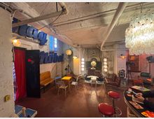 Gemütlicher Raum mit industriellem Look, weiß gestrichenen Backsteinwänden und sichtbaren Rohren an der Decke. Der Raum ist mit einer Mischung aus Vintage-Möbeln, bunten Stühlen, roten Barhockern und dekorativen Lichtelementen eingerichtet, einschließlich eines Kronleuchters und alter Filmbeleuchtung.