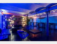 Ein moderner Lounge-Bereich mit dunklem Fußboden, Holzwand mit hängenden Pflanzen und Lichterketten sowie Sitzgelegenheiten aus schwarzen Lederbänken und Stühlen. Die Decke besteht aus Ziegeln, und die Beleuchtung setzt warme und blaue Farbakzente.