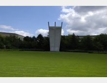 Eine große, graue Betonskulptur mit drei senkrechten Spitzen steht auf einer weitläufigen, gepflegten Rasenfläche, umgeben von Bäumen und Gebäuden im Hintergrund. Der Himmel ist teils bewölkt mit blauen Abschnitten.