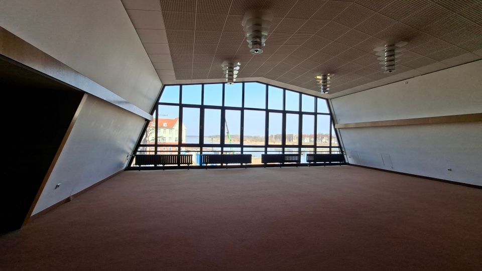 Großer, leerer Innenraum mit schrägen Wänden, Teppichboden und abgehängter Decke mit mehreren Deckenleuchten. Eine breite Fensterfront über die gesamte Stirnseite bietet Ausblick auf Gebäude und eine offene Außenfläche.