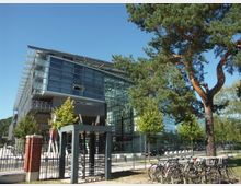 Modernes Bürogebäude mit Glasfassade und markanten, farbigen Stützpfeilern am Eingang. Im Vordergrund stehen Fahrräder an einem Abstellplatz, daneben ein hoher Baum, und der Himmel ist wolkenlos.