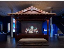 Eine Ausstellungsinstallation in einem Museum mit einer traditionellen japanischen Holzbühne, auf der eine Projektion von Trommlern und Tänzern zu sehen ist. Der Raum hat eine dunkle Beleuchtung, blaue Wände und Sitzbänke aus Holz vor der Bühne.
