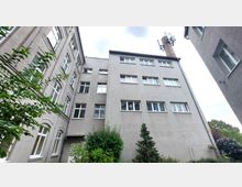 Mehrstöckiges Wohngebäude mit grauer Fassade und weißen Fensterrahmen, umgeben von einigen Bäumen und Sträuchern im Innenhof. Im Hintergrund ist ein Schornstein mit einer Mobilfunkantenne erkennbar.