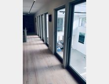 Flur mit hellem Holzboden und einer Reihe großer raumhoher Fenster auf der rechten Seite, durch die ein moderner Innenraum mit hohen Decken und Möbeln sichtbar ist. Links befinden sich Regale und eine dezente Beleuchtung an der Decke.