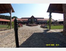 Ein gepflasterter Hof mit blühenden Beeten führt zu einem Gebäude mit rotem Ziegeldach und Giebel in der Mitte, flankiert von hölzernen Seitentrakten. Im Hintergrund ist eine Baumreihe sichtbar, und das Gelände ist von Rasenflächen umgeben.