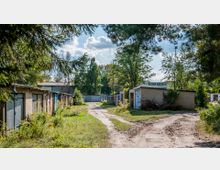 Reihe von Garagen mit alten Toren in einem überwucherten Gelände; ein unbefestigter Weg führt zwischen den Garagen hindurch, umgeben von Bäumen und Sträuchern bei sonnigem Wetter. Im Hintergrund ist weiteres Grün sowie ein größeres Gebäude sichtbar.