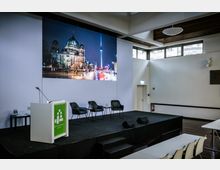 Moderner Veranstaltungsraum mit Bühne, bestehend aus einem Rednerpult, drei Stühlen und einem Beistelltisch mit Wassergläsern. Im Hintergrund hängt ein großes Bild mit einer nächtlichen Stadtansicht, das den Berliner Dom und den Fernsehturm zeigt.