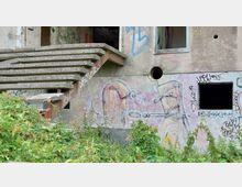 Außenansicht eines verlassenen Gebäudes mit Betonwänden, Graffiti und einem beschädigten Treppenaufgang. Im Vordergrund sind dichte Pflanzen, während die Wände des Gebäudes mit bunten Zeichnungen und Tags bedeckt sind.