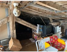 Dachboden mit sichtbarer Holzbalkenkonstruktion und niedriger Decke, ausgestattet mit verschiedenen Möbelstücken wie einem bunten Sofa, Stühlen, einem alten Röhrenfernseher und Körben. Im Hintergrund sind dunkle Bereiche, lagernde Gegenstände und ein Steinmauerwerk erkennbar.