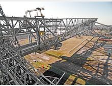 Industrielle Förderanlage mit einer großen Metallstruktur, die sich horizontal über eine offene Fläche mit Gras erstreckt. Im Hintergrund sind weitere industrielle Elemente und eine flache Landschaft zu sehen.