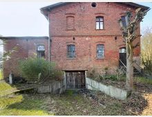 Backsteinbau mit Fenstern mit Rundbögen, einer Holztür im Untergeschoss und einem kleinen Treppenaufgang rechts. Vor dem Gebäude befinden sich ein bepflanzter Grünbereich und ein beschnittener Baum.