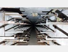 Rostiger, metallener Gang in einem engen, grauen Betonraum, mit symmetrisch angeordneten Metallhalterungen an den Seitenwänden und einem dunklen Fluchtpunkt in der Tiefe.