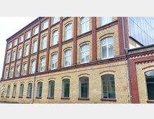 Mehrstöckiges Gebäude aus gelben und roten Backsteinen im traditionellen Baustil, mit großen, rechteckigen Fenstern, die durch rote Backsteinbögen eingerahmt sind. Im Vordergrund ist ein Straßenschild sichtbar.