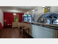 Kleine Bar mit weißer Theke und Holzstühlen in einem Innenraum. Im Hintergrund stehen Kühlschränke mit Flaschen, darüber hängen runde Lampen; die Wände sind rot und weiß gestrichen, teilweise mit dekorativen Elementen wie einem Kronleuchter und einem bemalten Schild versehen.