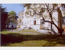 Großes, helles Gebäude im klassizistischen Stil mit Säulen am Eingangsbereich und mehreren Fenstern. Der Vordergrund zeigt eine Rasenfläche mit Bäumen, im Hintergrund spazieren mehrere Personen.