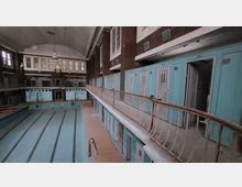 Innenraum eines alten Schwimmbads mit einem leeren, gefliesten Becken, umgeben von zweistöckigen Kabinen mit türkisfarbenen Türen. Die hohen Wände sind aus dunkelbraunem Mauerwerk, und durch große Fenster fällt gedämmtes Licht in den Raum.