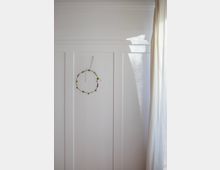 Weiße Wand mit vertikalen Paneelen, an der ein einfacher Kranz aus Draht mit kleinen grünen Blättern und roten Akzenten hängt. Rechts im Bild hängt ein heller Vorhang, durch den Sonnenlicht direkt auf die Wand fällt.