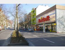 Straßenszene mit breiter Fahrbahn und Mittelstreifen, gesäumt von kahlen Bäumen. Rechts ein modernes Gebäude mit einer Supermarkt-Fassade und auffälligem "Reichelt"-Schriftzug; links farbenfrohe Mehrfamilienhäuser und parkende Autos.