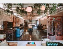 Großer, loftartiger Raum mit sichtbaren Backsteinwänden, einer langen Holztafel in der Mitte und einer offenen Küche im Hintergrund. Hängende Pflanzen, industriell gestaltete Lampen und Möbel im Vintage-Stil prägen das Ambiente.