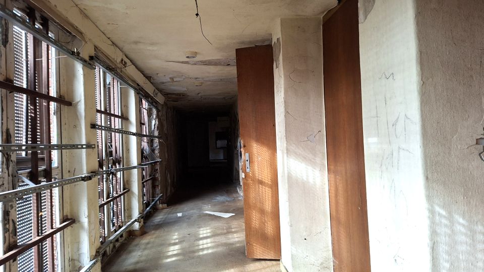 Langer, leerer Flur in einem heruntergekommenen Gebäude mit rissigen Wänden und Decke sowie Betonboden. Links verläuft eine Reihe hoher Fenster mit Metallgittern und Jalousien, rechts stehen mehrere braune Türen entlang des Gangs.