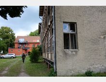 Zwei Gebäude in einer ländlichen Umgebung: Rechts ein älteres, teilweise verfallenes Gebäude mit verputzter Fassade und großen Fenstern, links im Hintergrund ein modernes, orangefarbenes Haus mit Balkon und roten Dachziegeln. Im Vordergrund eine Person, die über eine Wiese geht, mit parkendem Auto und Bäumen im Hintergrund.