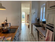 Schmale Küche in einem modernen Landhausstil mit Holzarbeitsplatten, Fliesenboden und weißen Wandfliesen. Im Hintergrund führt ein heller Flur mit Holzboden zu einer offenen Tür, durch die Sonnenlicht und ein Blick in den Garten sichtbar sind.