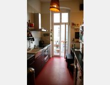 Schmale Küche mit roten Fußbodenfliesen, dunklen Unterschränken, weißem Spülbecken und moderner Kochstelle. Am Ende des Raums befindet sich eine große Glastür mit Blick auf einen kleinen Balkon mit Tisch und Stühlen.