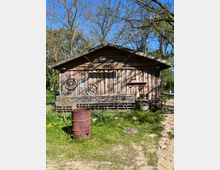 Holzhütte in ländlicher Umgebung mit rustikalem Aussehen, verziert mit alten Wagenrädern an der Fassade. Im Vordergrund steht eine rostige Metalltonne, umgeben von Gras und Wildblumen, während dahinter Bäume in einer natürlichen Kulisse sichtbar sind.