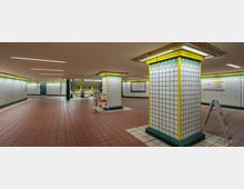 U-Bahn-Station mit gefliesten Säulen, die gelbe und grüne Dekorationselemente aufweisen, roten Bodenfliesen und hellen Deckenbeleuchtungen. Im Hintergrund befinden sich ein Treppenaufgang, Sitzbänke und Hinweisschilder.