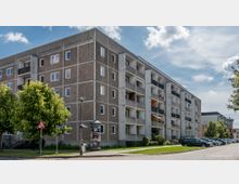 Mehrstöckiges Wohngebäude aus der Nachkriegszeit mit grauer Fassade und Balkonen auf jeder Etage. Im Vordergrund sind Bäume, ein Gehweg, geparkte Autos und eine Litfaßsäule zu sehen, bei klarem Himmel mit einigen Wolken.