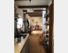 Langer, schmaler Wohnraum mit sichtbaren Holzbalken an der Decke. Im Vordergrund eine kleine Küche mit Holzarbeitsfläche und offenen Regalen, im Hintergrund ein Essbereich mit Tisch und Stühlen sowie dekorativen Bildern an der Wand.