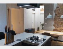 Moderne offene Küche mit Kochinsel, einschließlich eines Gaskochfelds und eines Messerblocks. Im Hintergrund sind eine Ziegel- und Putzwand, Esstisch mit Hängelampen sowie eine kleine Sitzbank und Türen zu angrenzenden Räumen zu sehen.