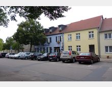 Straßenszene mit einer Reihe von geparkten Autos vor einem Gebäudeensemble mit verputzten Fassaden in Beige und Blau, auf dem mittleren Gebäude das Schild „Der Seehof“. Im Vordergrund eine Straße, im Hintergrund Bäume.