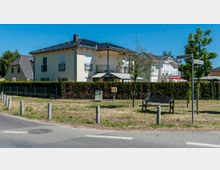 Straßenecke in einem Wohngebiet mit modernen, zweistöckigen Einfamilienhäusern und gepflegten Zäunen, einer kleinen Grünfläche mit Bank, Straßenlaternen und Straßenschildern mit den Namen „Pestalozzistraße“ und „Martin-Luther-Str.“. Im Hintergrund sind Bäume und blauer Himmel sichtbar.