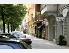 Straßenszene mit Bäumen und Autos entlang eines Bürgersteigs, gesäumt von Gebäuden mit Geschäften und Balkonen. Die Fassaden sind in hellen Farben gehalten, und es gibt Schilder sowie kleine Pflanzkübel vor den Geschäften.